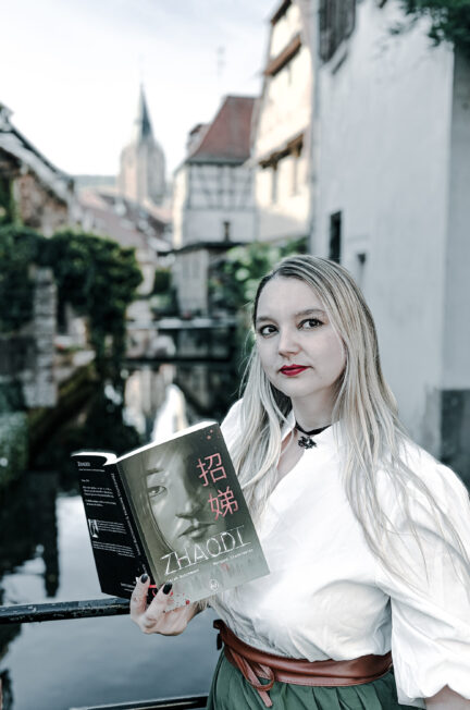 Photographie d’une femme blonde aux cheveux longs, vêtue d’une blouse blanche et d’une jupe verte, tenant ouvert le roman Zhaodi de Sarah Buschmann et Morgane Stankiewiez. Elle se tient près d’un cours d’eau, devant des maisons anciennes, avec un clocher visible à l’arrière-plan. La photographie accompagne une chronique littéraire du roman publié aux éditions Livr’S.