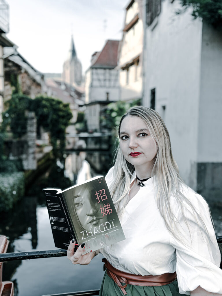 Photographie d’une femme blonde aux cheveux longs, vêtue d’une blouse blanche et d’une jupe verte, tenant ouvert le roman Zhaodi de Sarah Buschmann et Morgane Stankiewiez. Elle se tient près d’un cours d’eau, devant des maisons anciennes, avec un clocher visible à l’arrière-plan. La photographie accompagne une chronique littéraire du roman publié aux éditions Livr’S.