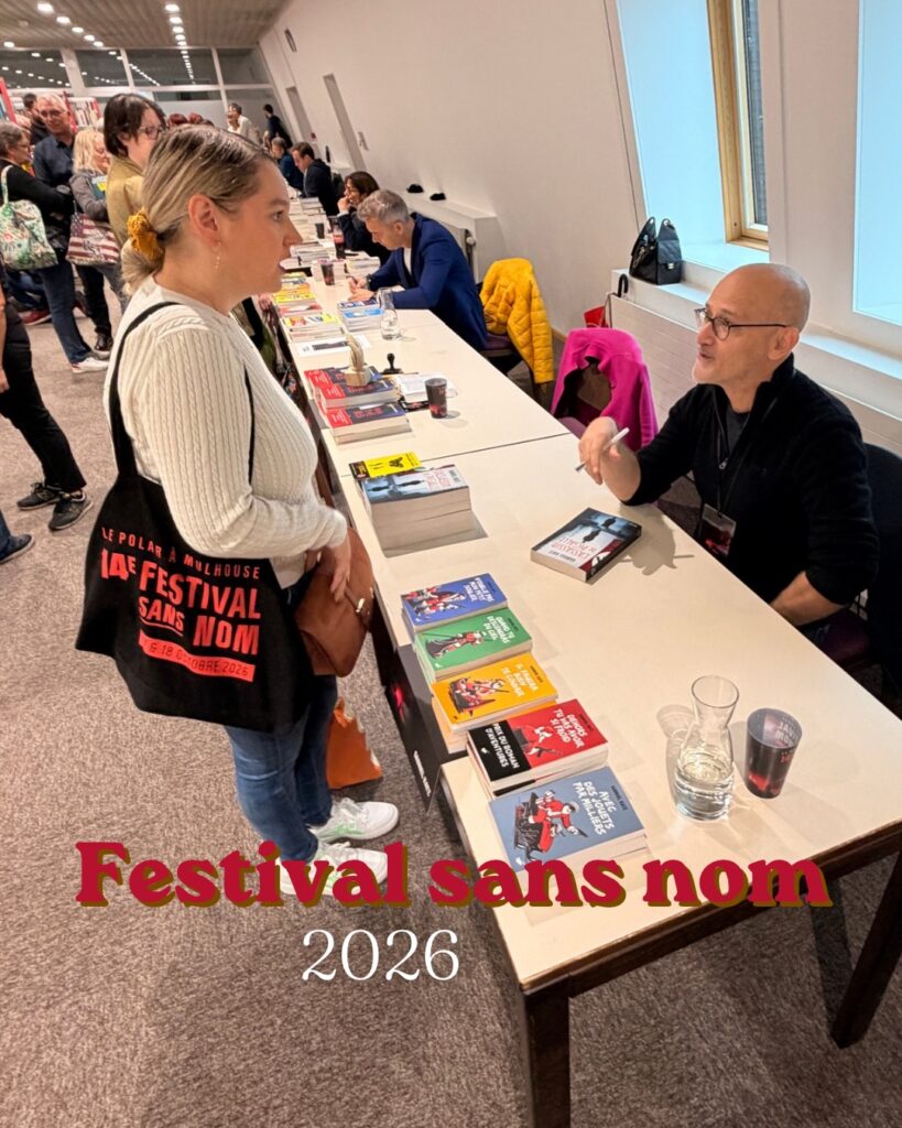 Photographie prise lors de la 14e édition du Festival Sans Nom, organisée à la bibliothèque Grand'Rue de Mulhouse en octobre 2026. On y découvre une discussion de Sorbet-Kiwi avec l'auteur Gabriel Katz, installé derrière une table de dédicaces, entouré de ses romans. D'autres auteurs et visiteurs sont visibles dans la salle. La photographie illustre l'ambiance conviviale de cette édition particulière du festival.